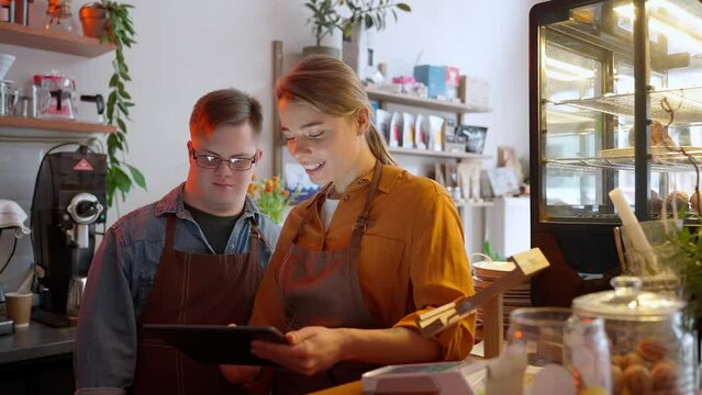Positive man barista with Down syndrome looking at tablet and talking with his girl colleague in a cafe