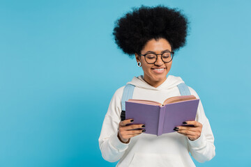 curly and stylish african american student in eyeglasses smiling while reading book isolated on blue.