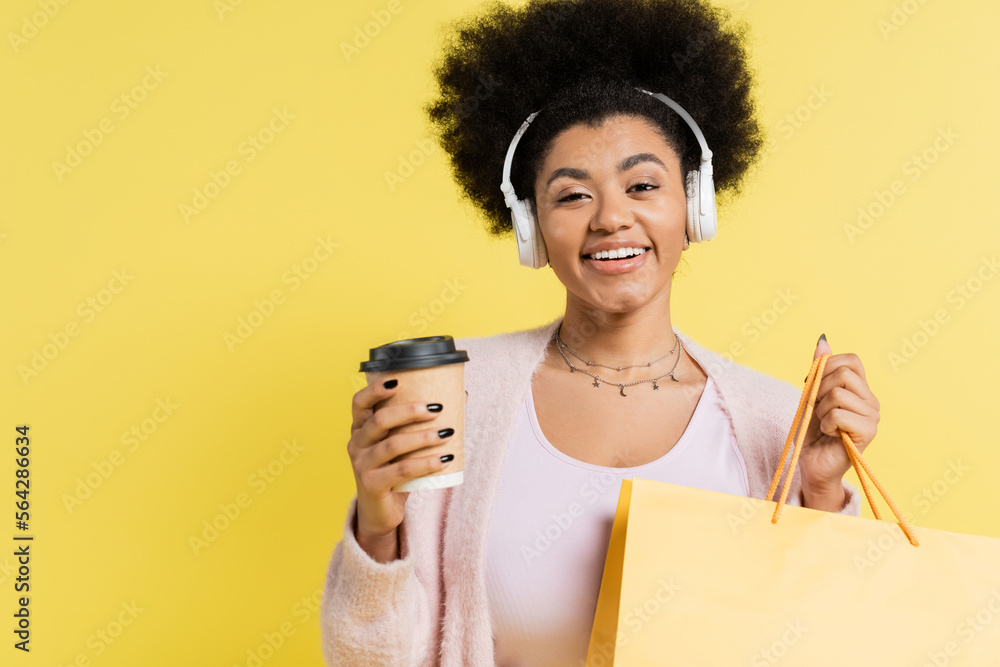 Sticker positive african american woman in wireless headphones standing with coffee to go and shopping bag isolated on yellow.