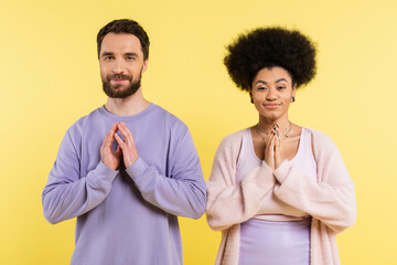 cunning interracial couple smiling at camera and showing please gesture isolated on yellow.