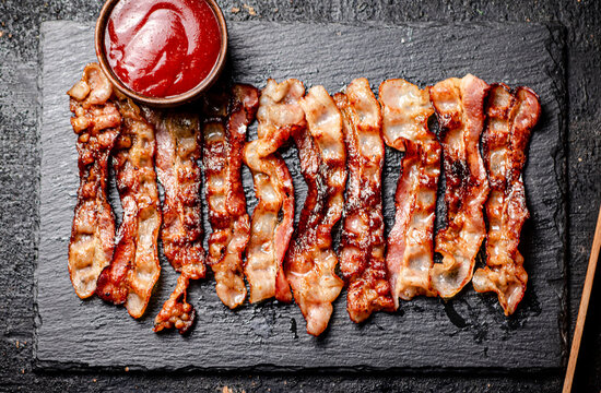 Strips of fried bacon on a stone board with tomato sauce. 