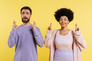 worried bearded man and optimistic african american woman crossing fingers isolated on yellow.
