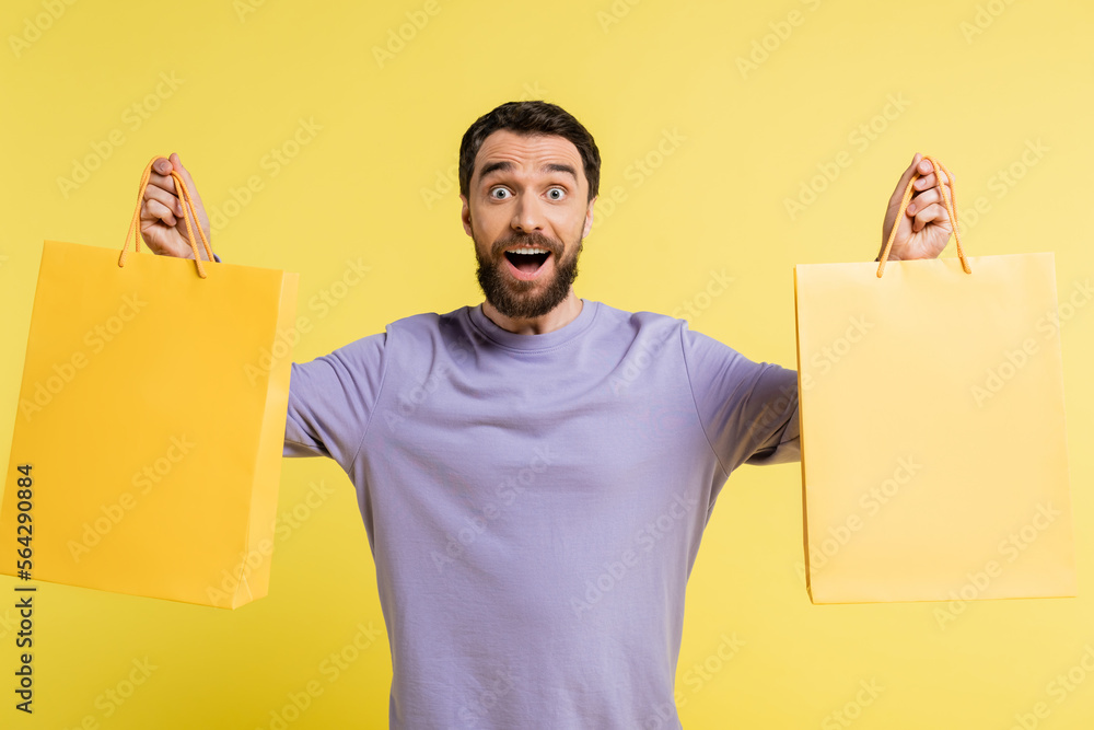 Canvas Prints amazed bearded man in purple pullover holding shopping bags and looking at camera isolated on yellow