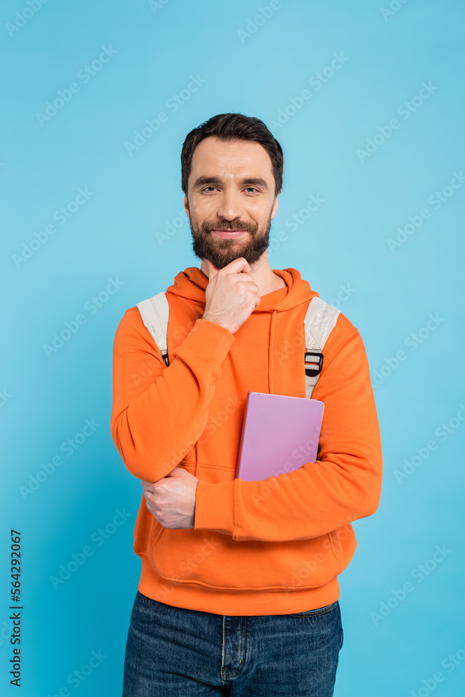 Wall mural smiling student with backpack and book touching beard and looking at camera isolated on blue. - Wall murals