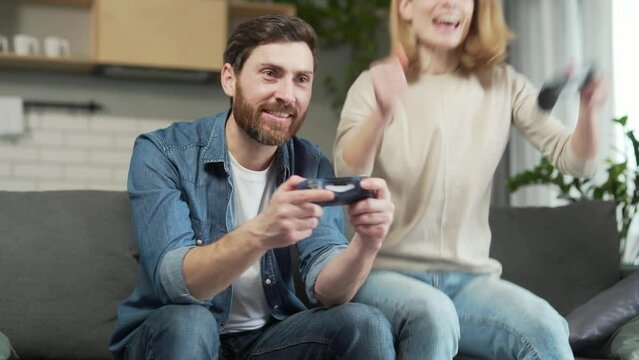 Happy adult man and woman playing with joysticks on play station exciting computer game while sitting on sofa in living room at home Сheerful couple laughs and dynamically moves to the beat of game