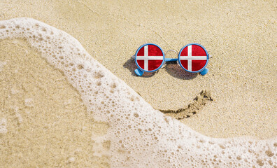 Fototapeta na wymiar Sunglasses with flag of Denmark on a sandy beach. Nearby is a sea lightning and a painted smile. travel and vacation concept