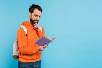 thoughtful and positive student with backpack reading book and touching beard isolated on blue.