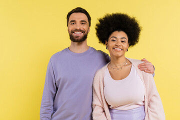 happy bearded man hugging shoulders of stylish african american woman while looking at camera isolated on yellow.