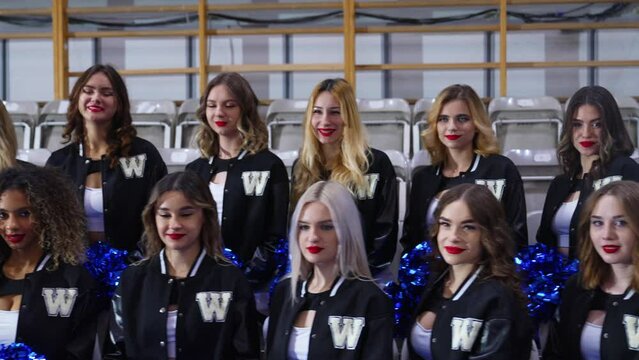 Front view of cheerleaders in black and white uniforms sitting on the stands in two rows while holding blue pom-poms. High quality 4k footage