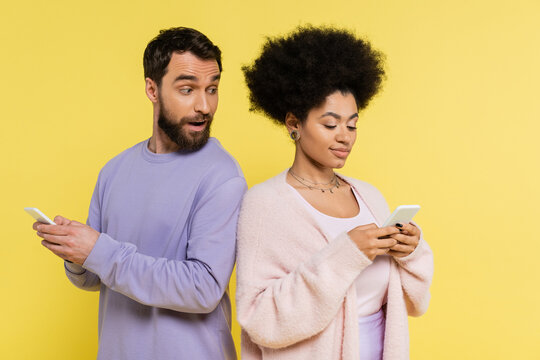 curious man with open mouth looking at african american woman messaging on smartphone isolated on yellow.
