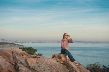 Naklejka na ściany i meble Young beautiful woman wearing eyeglasses looking on the sunlight sit on the rock over the sea