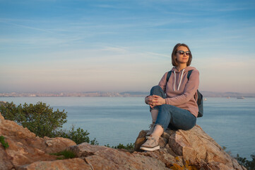 Fototapeta na wymiar Young beautiful woman wearing eyeglasses looking on the sunlight sit on the rock over the sea