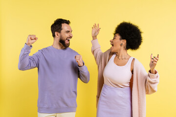 overjoyed multiethnic couple screaming and showing win gesture while looking at each other isolated on yellow.