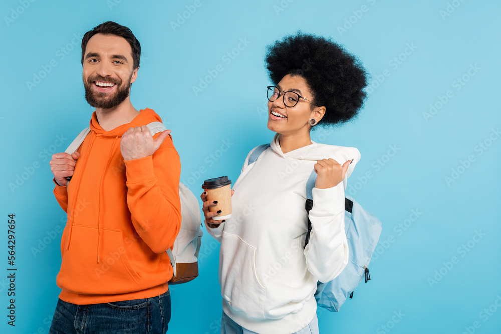 Wall mural cheerful interracial students with backpacks and coffee to go looking at camera and pointing with thumbs isolated on blue.