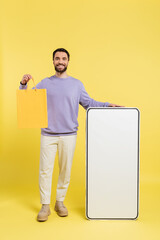 full length of cheerful bearded man standing with shopping bag and huge template of smartphone on grey background.