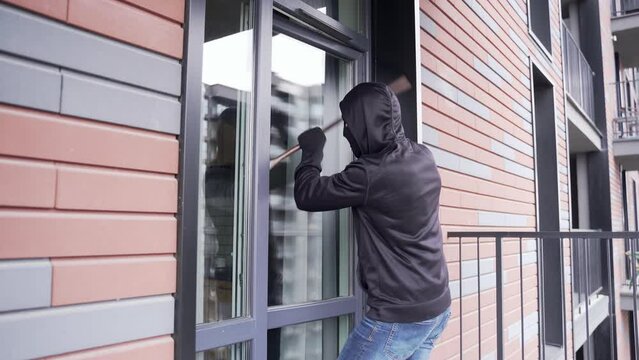 A thief sneaks into the apartment through the courtyard or balcony. A man in a black jacket and mask looks into the room through the glass door, breaks it open with a crowbar and enters the room