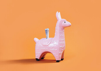coin money bank. Pink zine type toy alpaca llama with dollar note on orange background close up....