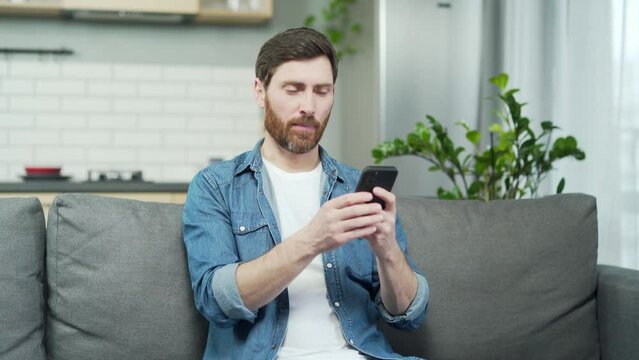 Single bearded man swiping through women profiles on dating app and like or dislike photos on smartphone in the living room Young guy looking for partner on social platform and waiting for a match