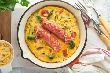 Omelette or frittata with two sausages, melted cheese, cherry tomatoes, broccoli and parsley on red skillet or iron pan on white old concrete background. Healthy breakfast food concept. Top view.