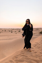 Beautiful mysterious woman in traditional arabic black long dress stands in the desert on sunset