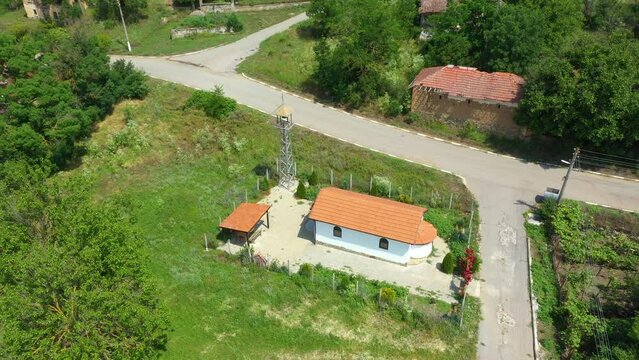 Aerial view of a church with bell tower in the heart of a small balkan village with a white house and a brown roof, a cloudy sky and a forest on the horizon.