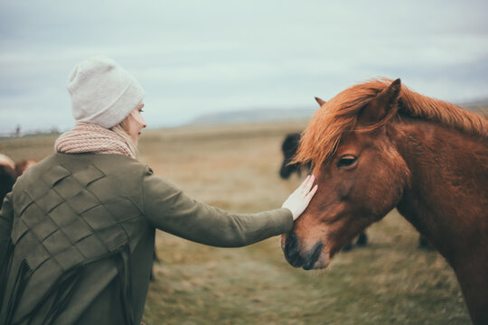 Stylish young woman walking on the nature. Tourist female pats the beautiful brown horse in the mountains in Iceland.