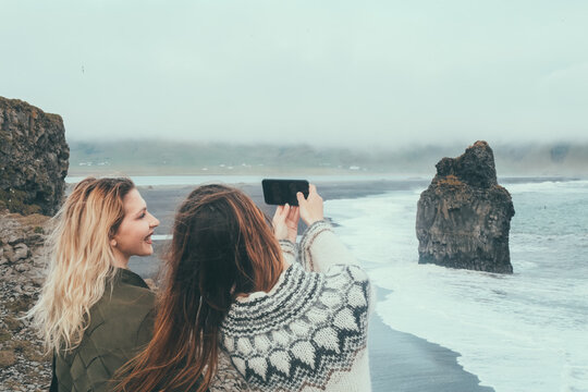 Beautiful friends taking selfie photo on smartphone on black beach in Iceland. Tourist female in lopapeysa holds camera.