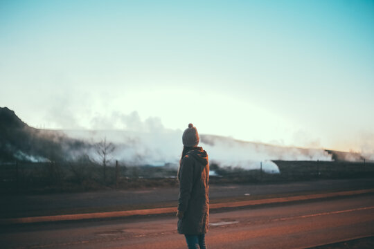 Back view of young beautiful woman standing in the evening near the hot springs in Iceland, looking at smoke.