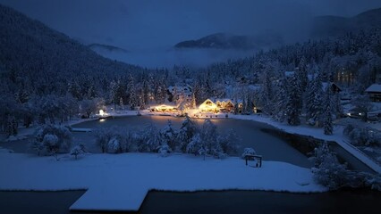 Aerial view of fairy forest in snow, frozen lake and house with lights at night in winter. Top view from drone of mountain village, pond, illumination, snowy pine trees, low clouds at dusk in Slovenia