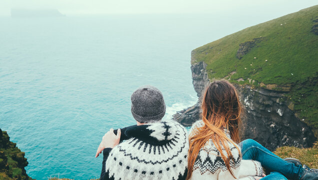 Traveling couple in lopapeysa sweaters sitting on the edge of the cliff and looking at the sea, enjoying the waves.
