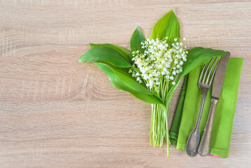 Bouquet of lilies of the valley, fork and knife on green napkin on wooden background; copy space