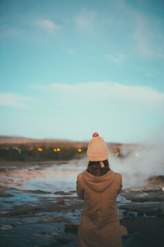 Back view of young stylish woman standing on the field and looking on the smoke from the hot springs in Iceland.