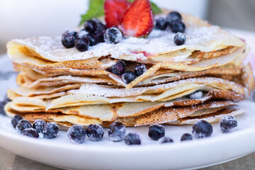 Pancakes with blueberries and mint leaf on top. A bunch of big homemade pancakes with forest fruits. A pile of flat thin cake in the background on a white plate.,wooden background
