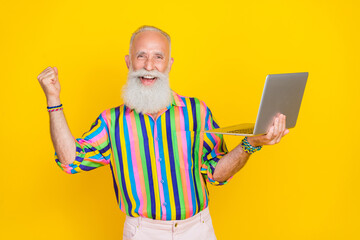 Portrait of positive successful old man with long beard wear colorful shirt hold laptop clenching fist isolated on yellow color background