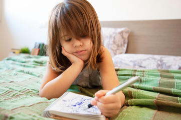 Little cute preschool girl lying on the bed in bedroom at home, she's bored and scribbling with blue pen on the paper notebook. Childhood concept. Leisure activity indoors. Getting bored at home.