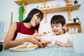 Happy little young boy and his mother making an easy breakfast together in a kitchen with a breads and chocolate jam. Mother and son playing together while making a breakfast.