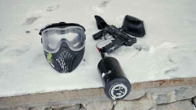 Slow motion video of a paintball mask and paintball guns lying on the snow. The camera moves smoothly close-up