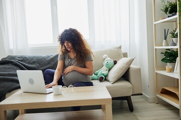 A pregnant woman sits at home on the sofa with a laptop and talks on video chat, blogger, freelancer works online. Home clothes, lifestyle of a pregnant woman, preparation for childbirth