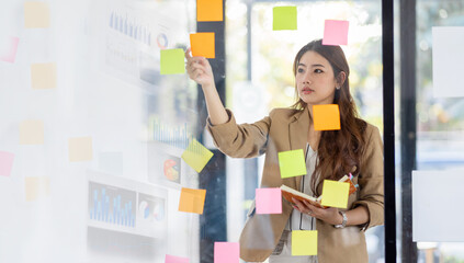 Fototapeta na wymiar Young Business Asian woman using sticky notes and graph chart in glass wall analyzing strategy business plan to development grow to success