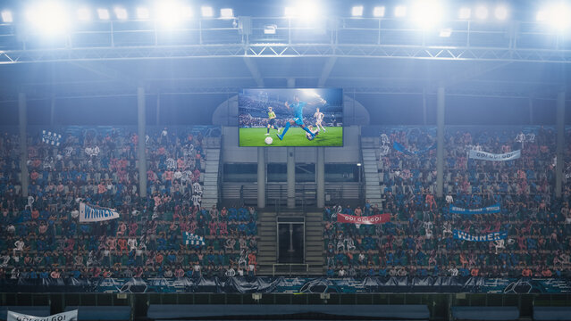 Football Soccer Championship Stadium Match, Scoreboard Screen Showing Repeat of the Goal. Crowd of Fans Cheering, Screaming, Having Fun. Sport Channel Television Advertising Concept. Wide Shot.