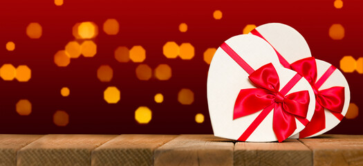 Banner with copy space. Valentine's day background with white heart shaped present boxes with red ribbon and bow on wooden surface table with blurred lights on red background - Powered by Adobe