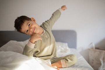 Cheerful energy cute little boy sits on bed, woke up and raises his hand up, stretching body in...