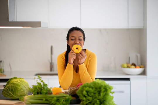 Cheerful hungry millennial african american lady in casual hold donut, enjoy aroma at table with organic vegetables