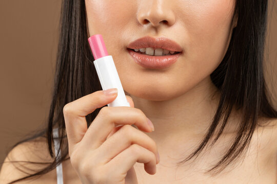 Cropped image of young asian lady holding pink lipstick in hand near face, brown background, studio shot, closeup