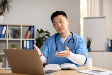 Serious mature chinese therapist in uniform look at computer, talking, gesturing at table in clinic