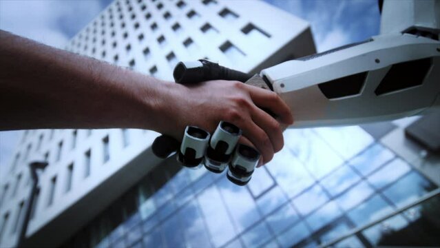 Close-up footage of robot and human shaking hands on blurred background of beautiful glass business center in city. Daytime. Cooperation. Artificial intelligence concept. Future