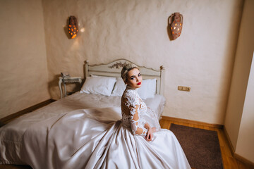 A beautiful, young blonde bride in a white long lace dress is sitting on a sofa, a bed in a room, a bedroom. Wedding photography, portrait.