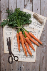 Mazzo di carote con foglie legate sopra fogli di spartito e altri elementi sparsi sul tavolo di legno © profumodarancia