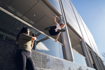 blond caucasian boy doing parkour jumping a fence in the city and a girl records him with the...