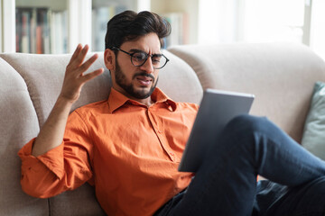 Angry middle eastern man sitting on couch, using digital pad